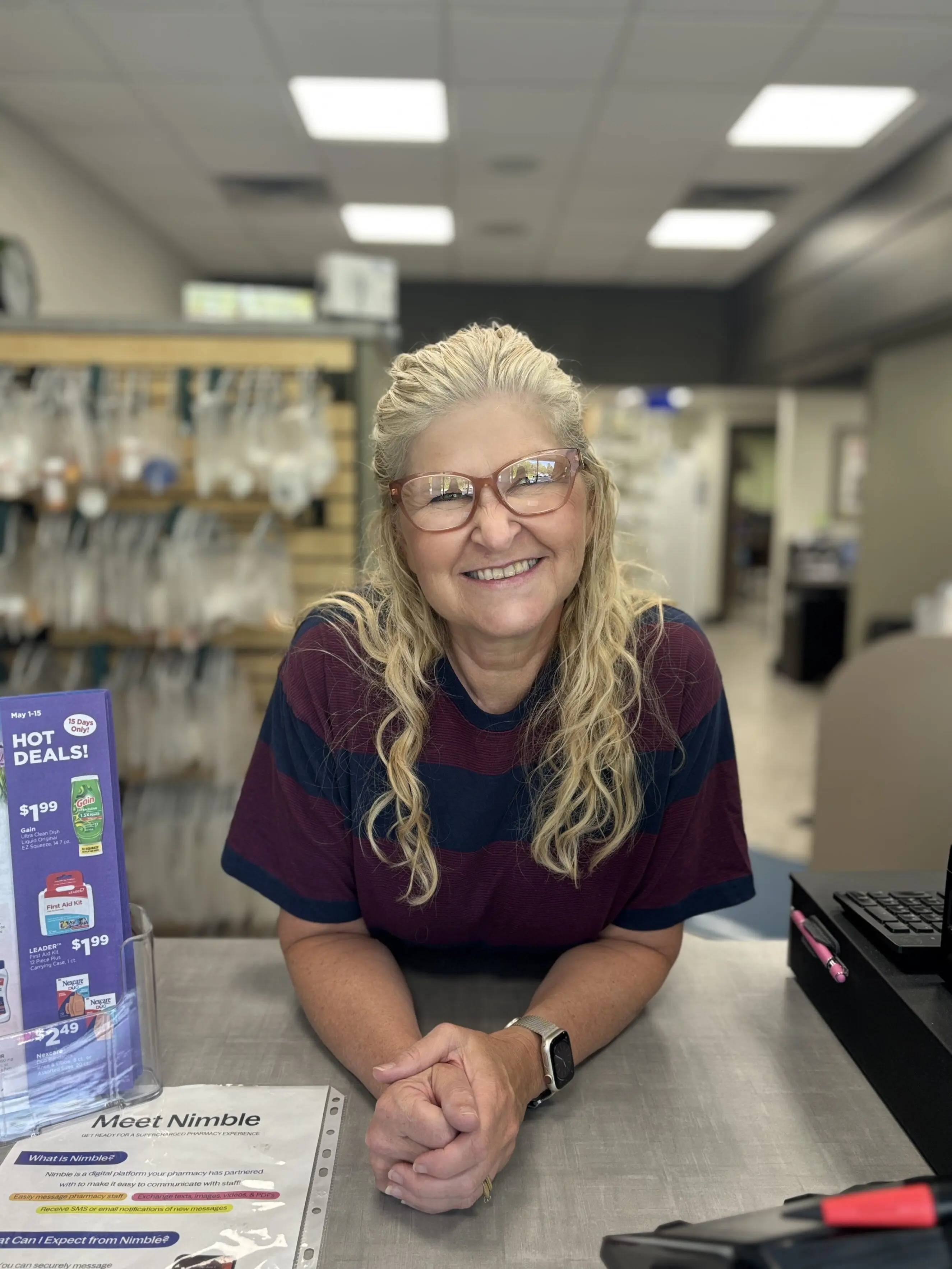 Female pharmacy staff member leaning on counter with folded hands, wearing a striped shirt and smartwatch, pharmacy shelves and promotional display for "Hot Deals" visible behind and to the left, counter paperwork titled "Meet Nimble" and a register with pens to the right — conveying a friendly, personalized neighborhood pharmacy setting focused on quick service and patient care